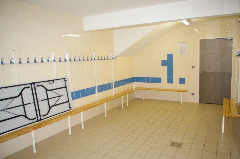 Vestiaire vide aux murs carrelés crème et accents bleus, bancs en bois, patères et table pliante